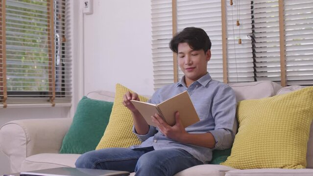 At home, a handsome young Asian businessman in casual clothing reads a book on the couch.