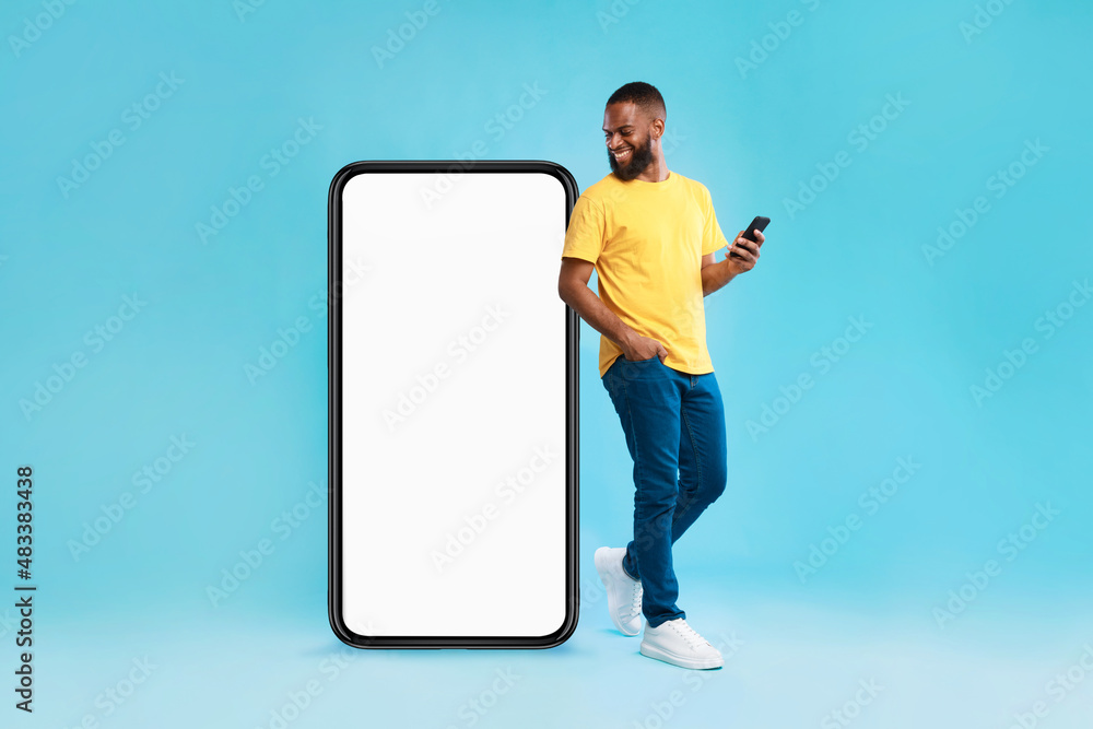 Aufkleber cheery millennial black guy with mobile device looking at giant smartphone, mockup for mobile app on