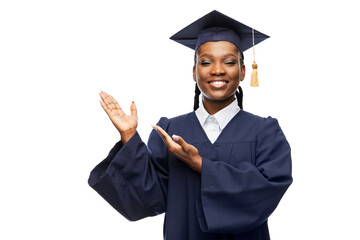 education, graduation and people concept - happy graduate student woman in mortarboard and bachelor gown showing something imaginary over white background