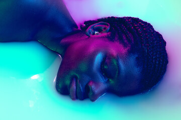 Profile view of female face of african girl in milk bath with soft white glowi in neon light. Modern neoned colors, foam. Beauty, fashion, style, skincare concept.
