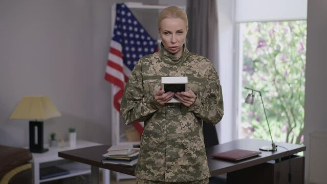 Frustrated lonely woman in military uniform admiring photo frame standing indoors with USA flag at background. Portrait of depressed sad young female recruit missing family. Defense industry lifestyle