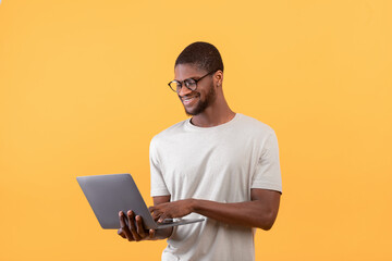 Freelance career. Positive african american guy using laptop and smiling, standing over yellow...
