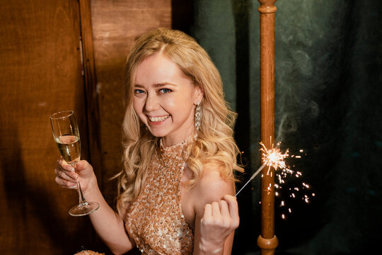 Cheerful young blond woman holding champagne flute and sparkler at New Year's party