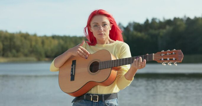 A cheerful girl with pink hair and glasses is standing near the lake, holding a guitar in hands, playing and singing favorite songs, fooling around, dancing