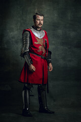 Vintage style portrait of brutal seriuos man, medieval warrior or knight with dirty wounded face holding sword isolated over dark background. Comparison of eras