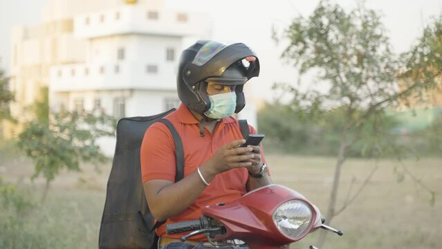 Young indian delivary boy with medical face mask searching adress on mobile phone using map or navigation - concept of technology , delivery service during coronavirus or covid-19 pandemic