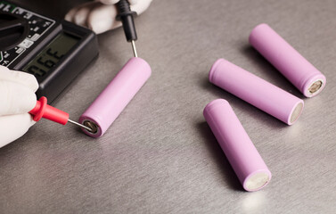 Multimeter red and black poles in the hands of a man checking voltage and amps, pink 18650 batteries, gray background