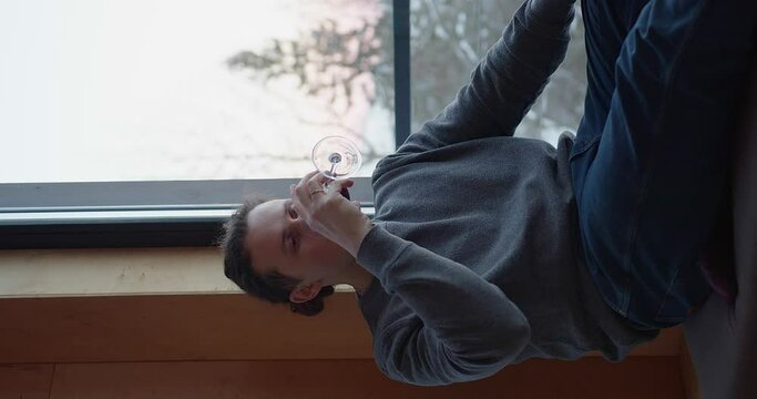 man with tied hair in a gray sweater sits on the windowsill in a beautiful room and drinks red wine, looking at the glass and then at the beautiful mountains outside the window. vertical video.