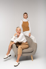 Fototapeta na wymiar full length of joyful young woman near smiling mother sitting in armchair on grey