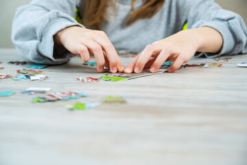 The child collects a puzzle. Elements of the puzzle are laid out on a wood table background. Children's hands during the game. Home leisure.