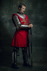 Portrait of brutal seriuos man, medieval warrior or knight with dirty wounded face holding sword isolated over dark background. Comparison of eras