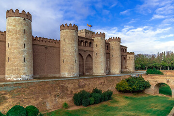 Zaragoza, Spain - November 29, 2021: Medieval stone facade with towers of Aljaferia Palace in Saragossa - outside view. Ancient fortress, moat around with green grass and bushes and arch bridge
