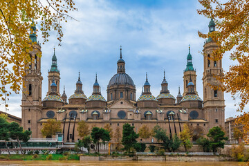 Fototapeta na wymiar Spanish Cathedral Basilica Nuestra Senora del Pilar among golden autumn trees on the embankment of the river Ebro with yellow water in Zaragoza. Beautiful architectural background with copy space