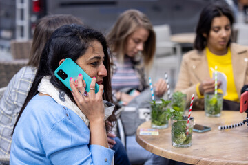Close up portrait of Indian young pretty joyful brunette woman sitting at a table outside of a cafe or pub in the city with female friends enjoying a cocktail in the sun and speaking on cellphone
