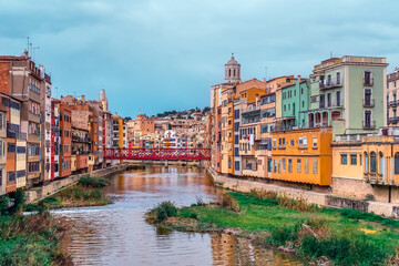 Fototapeta na wymiar Panorama of Girona with red metal lattice Eiffel Bridge over the river Onyar, Spain. Catalan old town with colorful buildings on the shoreline of the stream and their reflections in the water