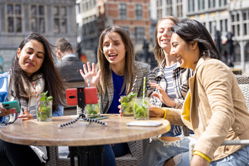 Four multi ethnic female friends, tourists or students sitting at a cafe terrace in the city center using mobile phone for a video call. High quality photo