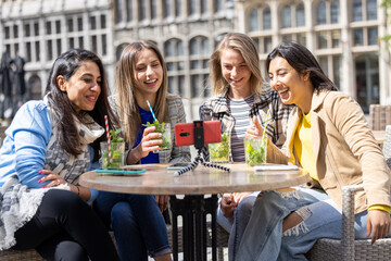 Antwerp, Belgium, May 21, 2021, four mixed race female tourists or students sitting outside in the old city center at a cafe terrace holding a video call using mobile phone. High quality photo