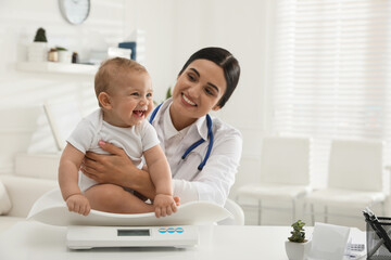 Young pediatrician weighting cute little baby in clinic. Space for text - obrazy, fototapety, plakaty