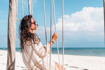 young hispanic woman with sunglasses and kimono sitting in a hammock looking at the caribbean sea