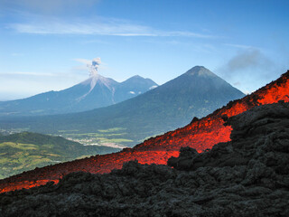 Volcanic river flowing between the solidified lava in the Pacaya volcano in Guatemala with an explosion of the Fuego volcano in the background with fumarole