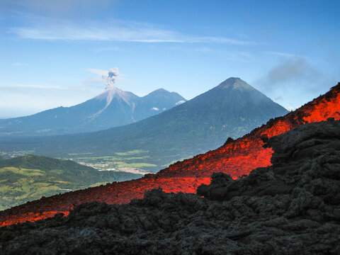Volcanic river flowing between the solidified lava in the Pacaya volcano in Guatemala with an explosion of the Fuego volcano in the background with fumarole