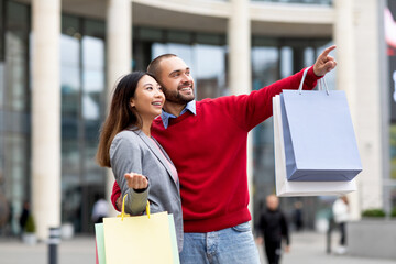 Positive diverse couple walking along street with paper bags, pointing at new store or advertisement, shopping together