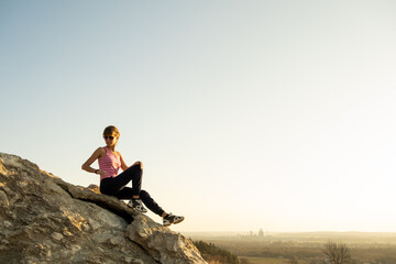 Naklejka na ściany i meble Woman hiker sitting on a steep big rock enjoying warm summer day. Young female climber resting during sports activity in nature. Active recreation in nature concept.