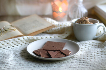 Plate with almond chocolate bar, mug with chocolate chip cookies, open book, reading glasses, soft blanket and lit candles. Hygge at home. Selective focus.