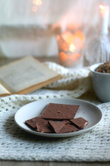 Plate with almond chocolate bar, mug with chocolate chip cookies, open book, reading glasses, soft blanket and lit candles. Hygge at home. Selective focus.