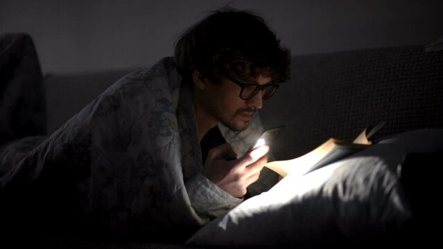 Man reads a book with a flashlight under the covers at night.