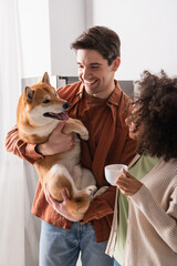 joyful man holding shiba inu dog near african american girlfriend with coffee cup