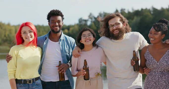 A group of students of different nationalities meet on lake during summer vacations, embrace each other standing on beach drink beer from glass bottles celebrate passing exams have fun together