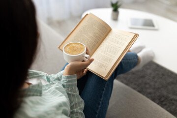 Obraz premium Unrecognizable Indian woman reading book, drinking fesh cappuccino at home. Hobbies and pastimes concept