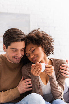 Man hugging african american girlfriend with cup of coffee at home