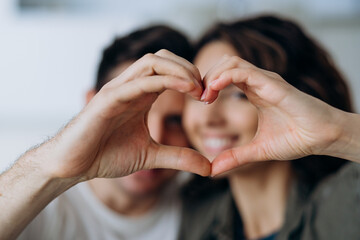 Couple in love hugging and showing a heart shape with their fingers and looking through it at the camera while sitting on the sofa in the living room.