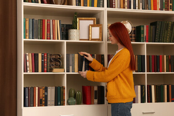 Beautiful young woman taking book from shelf in home library