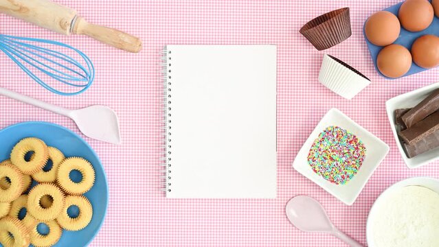 Baking ingredients and cookies in plate appear on pastel pink background with empty notebook for recipes. Stop motion flat lay