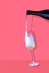 champagne is poured from a bottle into a glass on a pink background, with a shadow
