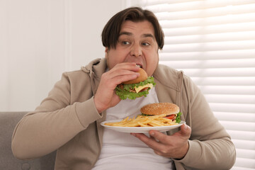 Overweight man eating tasty burger on sofa at home