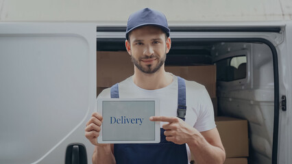 Smiling delivery man pointing with digital tablet with lettering near auto outdoors