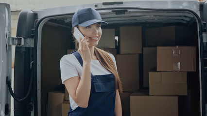 Smiling delivery woman talking on smartphone near cardboard boxes in car outdoors
