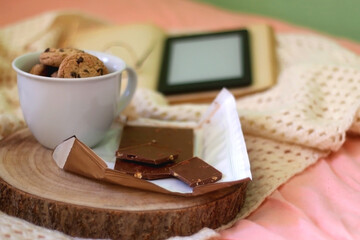 Mug of chocolate chip cookies, bar of chocolate, open book, reading glasses and e-reader on a bed. Selective focus.