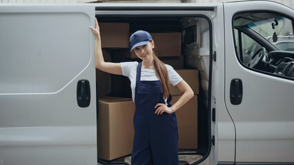 Smiling courier in overalls standing near cardboard boxes in car outdoors