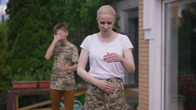 Stressed military mother looking at camera as angry teenage son shouting gesturing in slow motion at background. Portrait of depressed devastated beautiful Caucasian woman posing outdoors