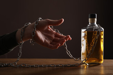 Alcohol addiction. Man chained with bottle of whiskey at wooden table, closeup - obrazy, fototapety, plakaty
