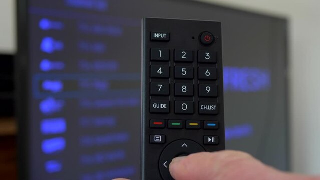 Closeup shot of a man's hand holding and pressing buttons on a remote control while facing a TV, to change the channel.