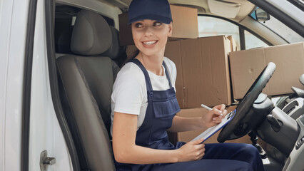 Cheerful delivery woman in uniform writing on clipboard near carton boxes in car