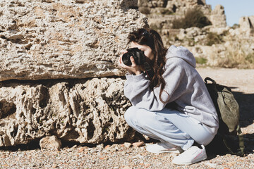 Portrait of young woman with camera. Sitting tourist taking photo of ancient ruins on sunny day. Blurred background.