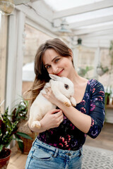 Young woman holding a white rabbit in her arms while standing in a home flower greenhouse