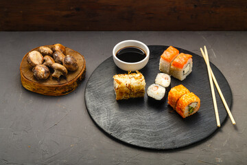 Assorted rolls on a round board next to sticks, soy sauce and baked champignons on a dark background.